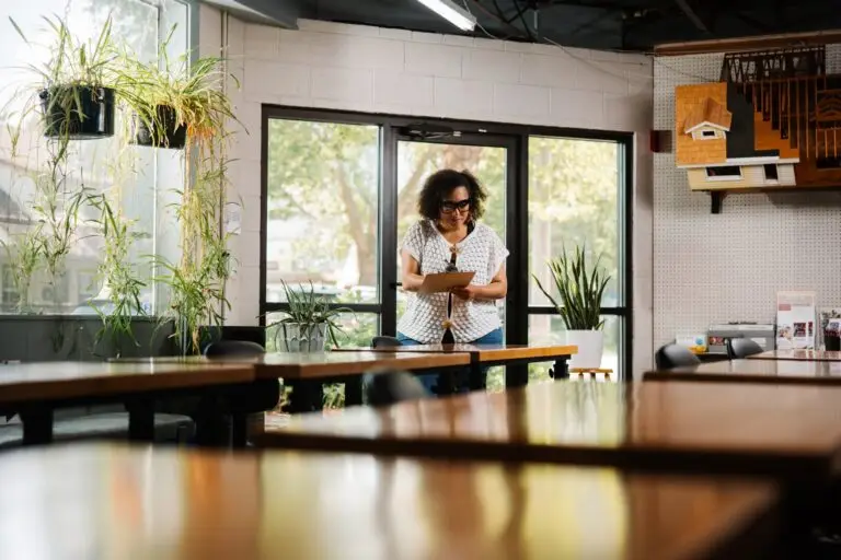 A woman holding a clipboard, standing in a room full of wooden desks.